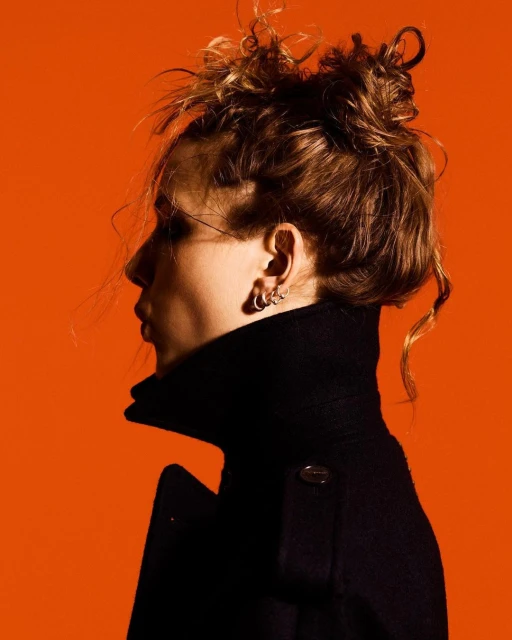 Profile portrait of a young woman with an updo wearing a black coat against a vivid orange background in a studio setting.