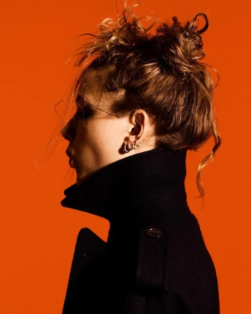 Profile portrait of a young woman with an updo wearing a black coat against a vivid orange background in a studio setting.