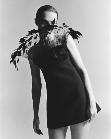 Fashion photo in black and white of a model in a black mini dress and feathered collar against a solid backdrop in a studio.