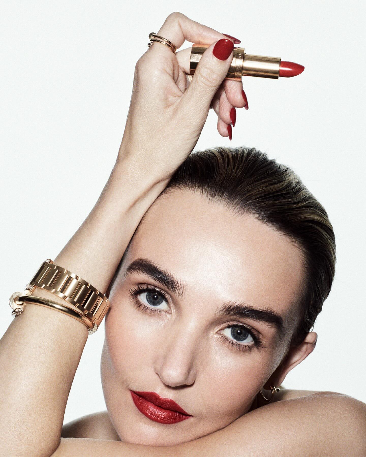 Close-up of a woman with red lipstick and manicured nails holding a gold tube of red lipstick against a white background.