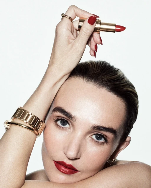 Close-up of a woman with red lipstick and manicured nails holding a gold tube of red lipstick against a white background.