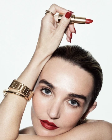 Close-up of a woman with red lipstick and manicured nails holding a gold tube of red lipstick against a white background.