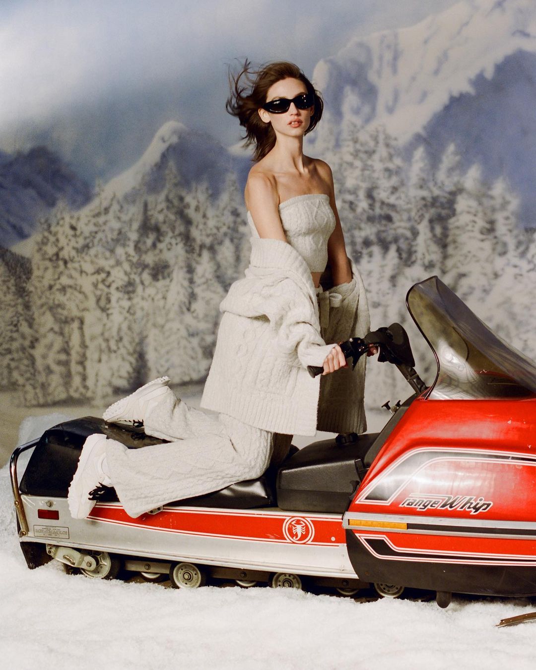 Woman in knitted outfit on a snowmobile against a backdrop of snowy mountains and trees, wearing sunglasses and looking forward.