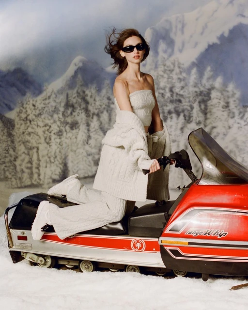 Woman in knitted outfit on a snowmobile against a backdrop of snowy mountains and trees, wearing sunglasses and looking forward.