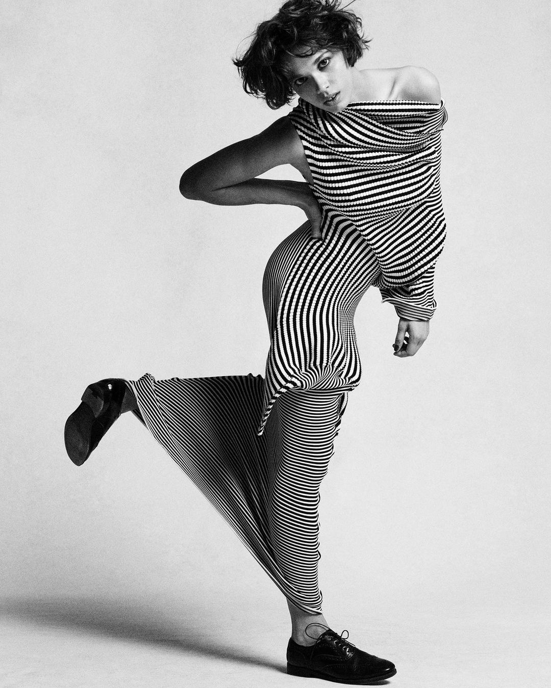 Woman in black and white striped dress and black oxfords poses in a studio with one leg raised for a fashion editorial shot.
