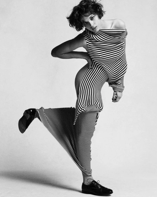 Woman in black and white striped dress and black oxfords poses in a studio with one leg raised for a fashion editorial shot.
