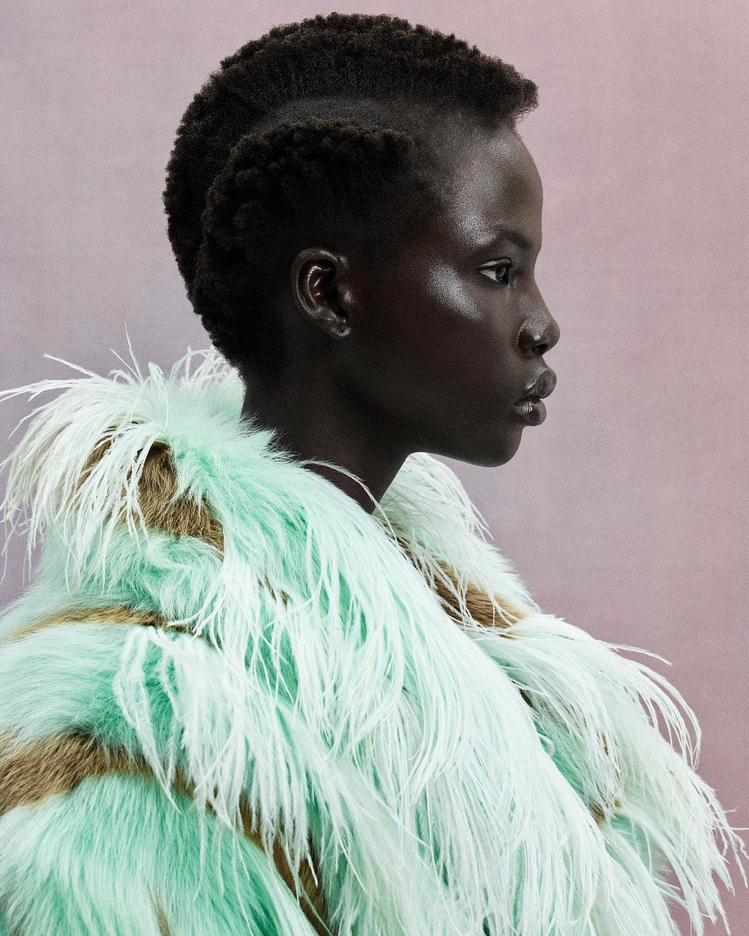 Profile portrait of a dark-skinned woman with a shaved head and an avant-garde feathered coat against a soft pink backdrop in a photo studio.
