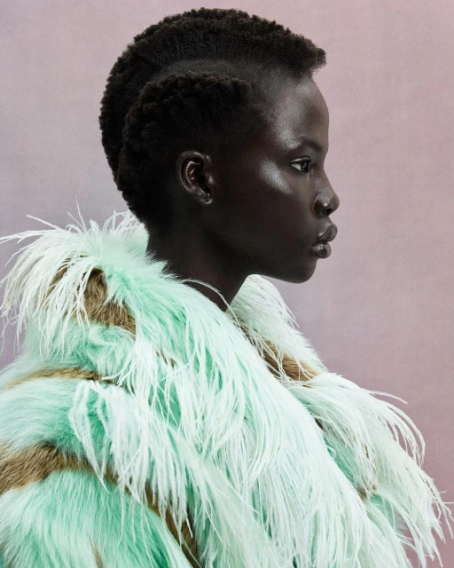Profile portrait of a dark-skinned woman with a shaved head and an avant-garde feathered coat against a soft pink backdrop in a photo studio.