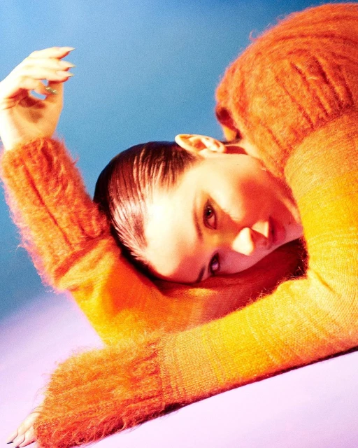 Portrait of a light-skinned female model in a fur-like orange sweater, lying down and looking at the camera on a blue and purple background.
