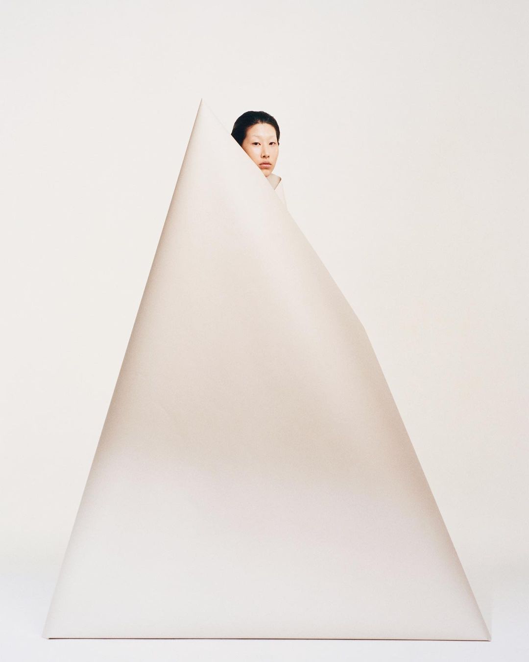 A woman with dark hair peeks out from behind a large, geometric, triangular shape against a plain white background in a studio setting.