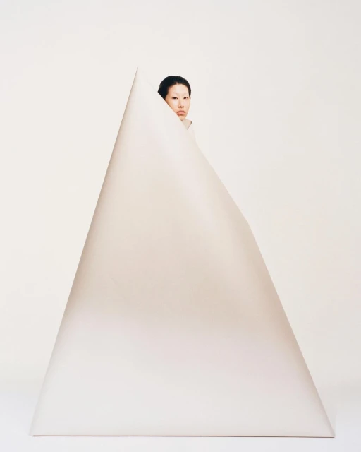 A woman with dark hair peeks out from behind a large, geometric, triangular shape against a plain white background in a studio setting.