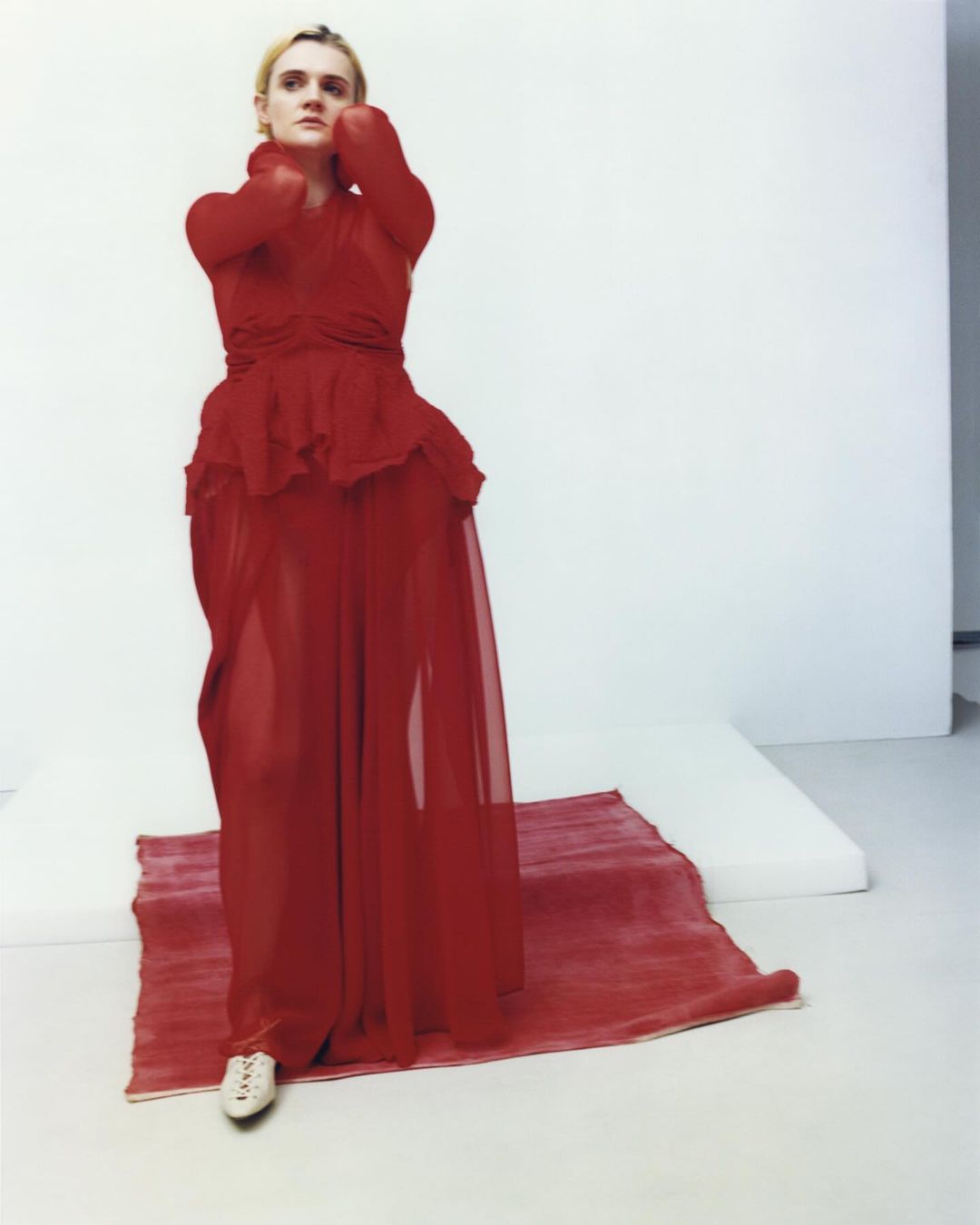 A fair-skinned, adult woman in a red, semi-sheer dress stands on a red rug over a white surface, with her hands at her face in a photo studio.
