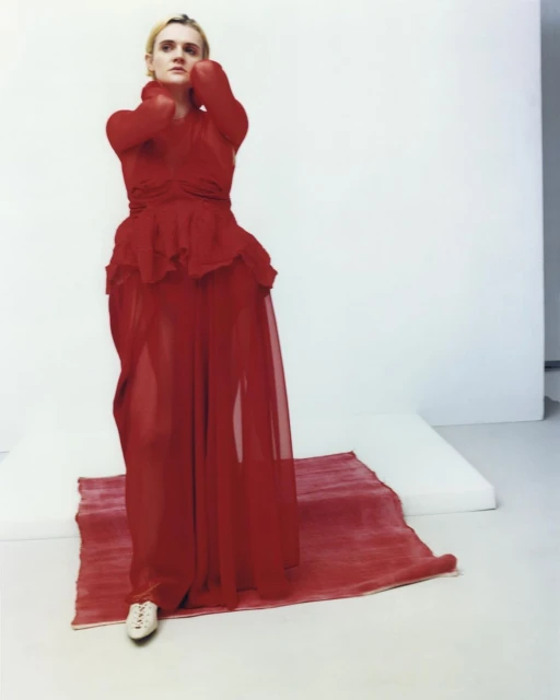 A fair-skinned, adult woman in a red, semi-sheer dress stands on a red rug over a white surface, with her hands at her face in a photo studio.