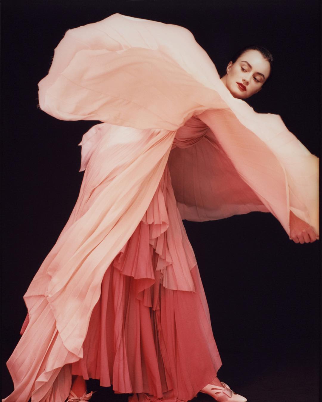 Fashion shot of a woman in a pink dress with flowing fabric on a dark background, dramatic pose and elegant style.