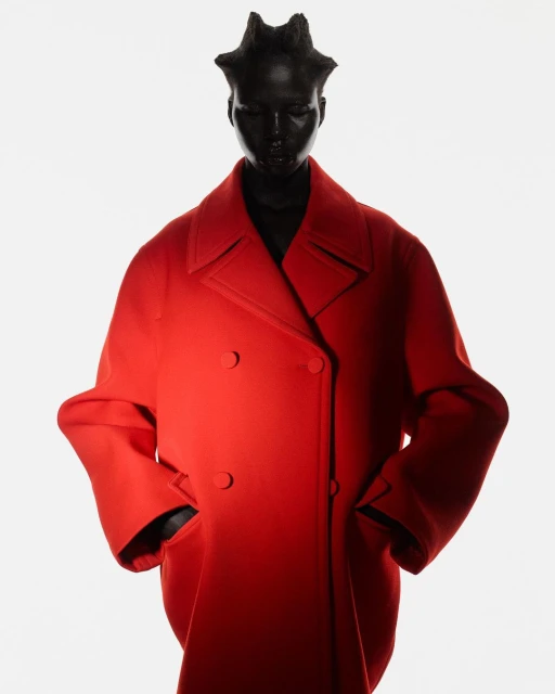 Fashion shot of a mannequin wearing a red overcoat against a white background in a studio setup. The red coat is the dominant subject.