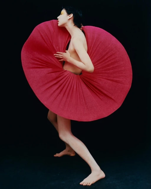 A striking fashion portrait of a model in a red sculptural garment against a black background, shot in a photo studio with vivid makeup.