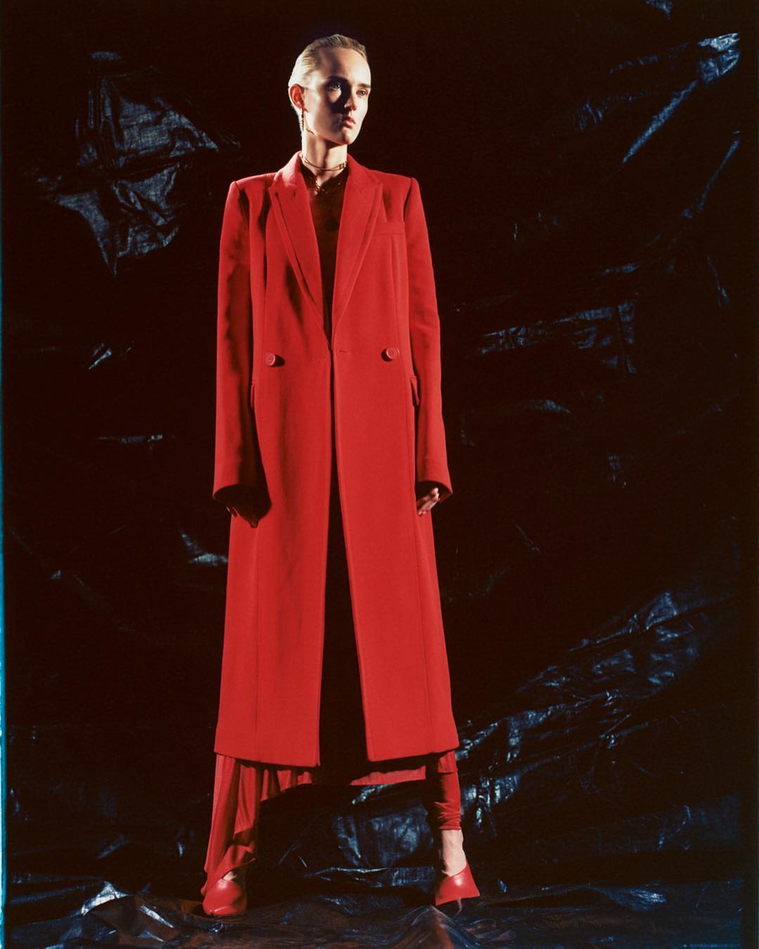 Fashionable adult woman wearing a long red coat and red shoes stands on a black textured surface in a studio setting with dark background.