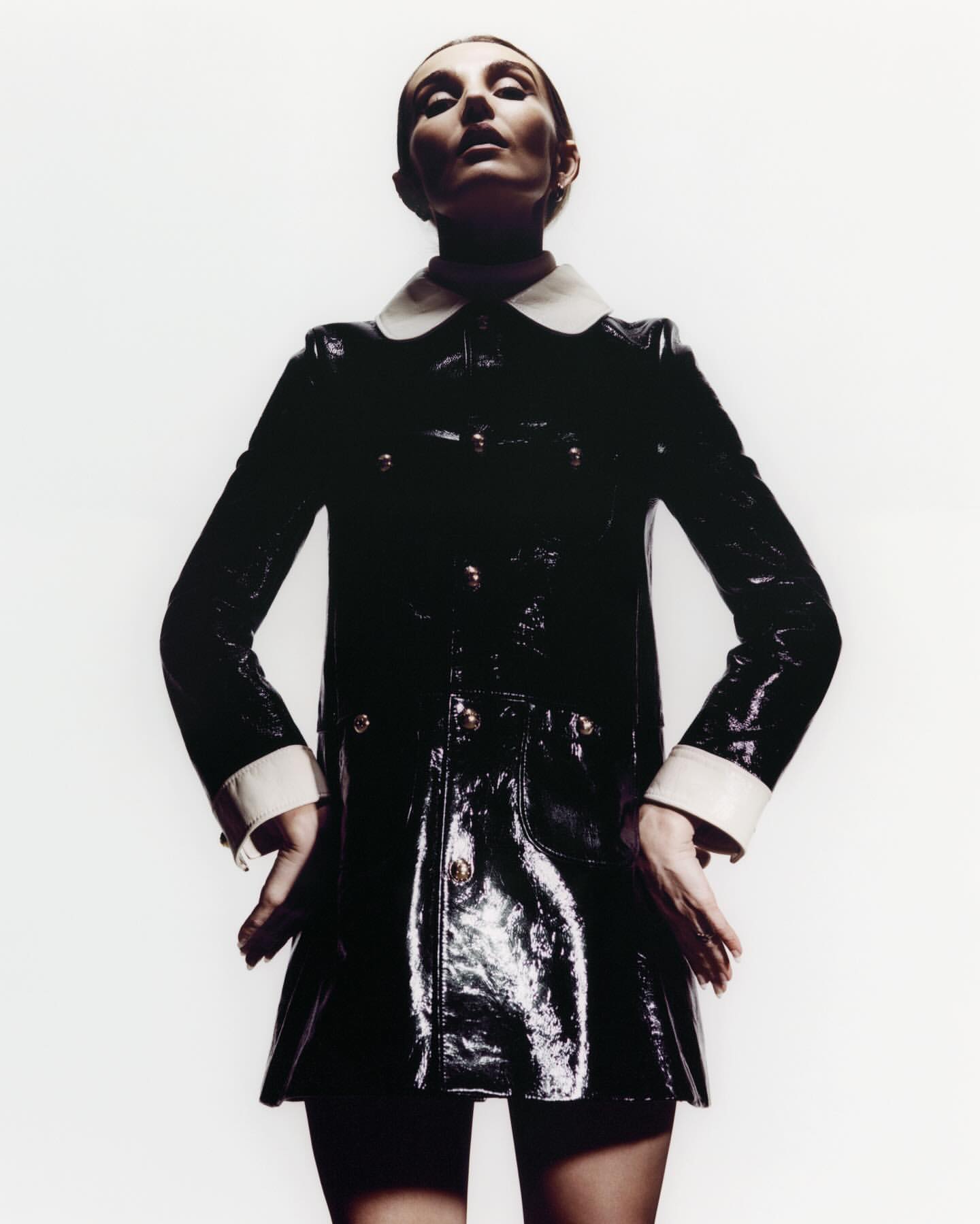 Fashionable woman posing in a glossy black coat with white collar and cuffs against a bright white backdrop, looking up at the camera.