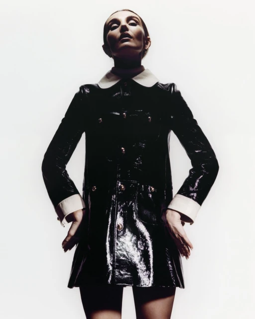 Fashionable woman posing in a glossy black coat with white collar and cuffs against a bright white backdrop, looking up at the camera.