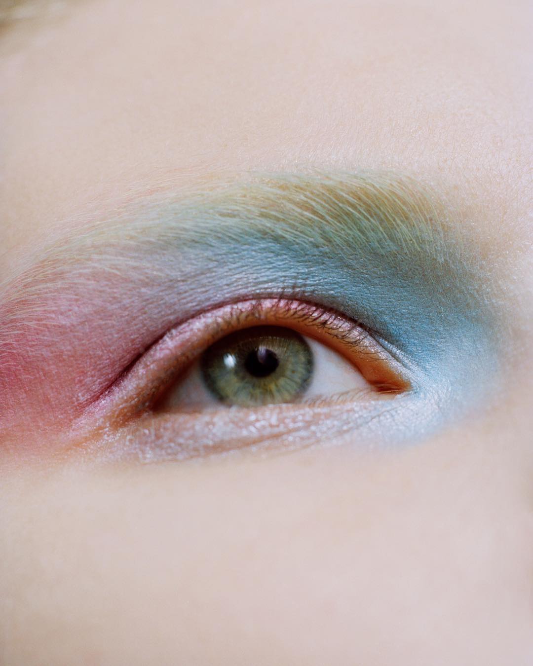 Close-up portrait of an adult woman's eye with creative, colorful eyeshadow in shades of pink, blue, and green, set against fair skin.