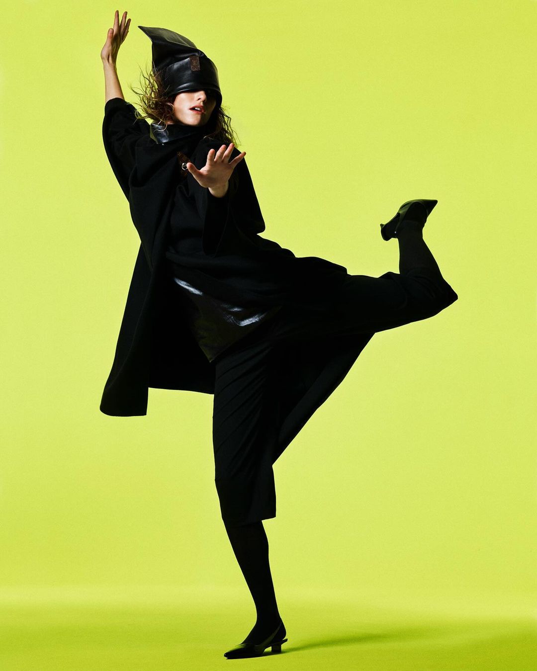 Model in black coat and pointed leather hat posing dynamically against a vibrant lime green background