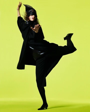 Model in black coat and pointed leather hat posing dynamically against a vibrant lime green background