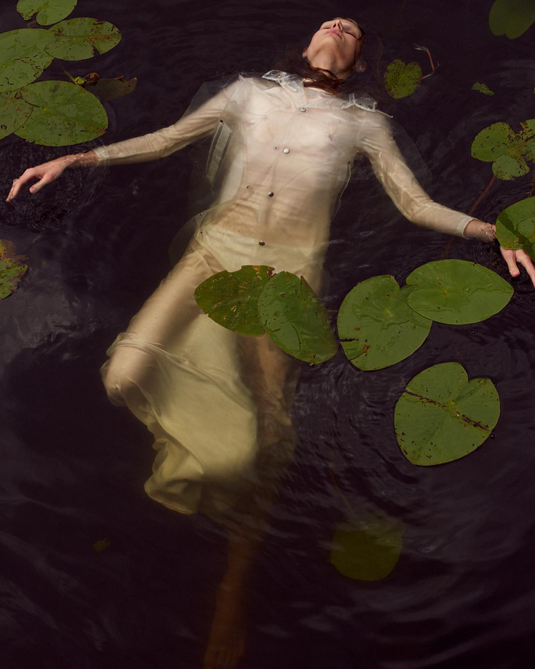 Woman in a sheer white dress floats serenely in dark water surrounded by green lily pads in an artistic, ethereal outdoor setting.