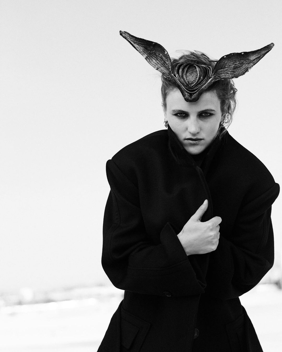 Model wearing a black coat and sculptural horned headdress against an overcast sky background.