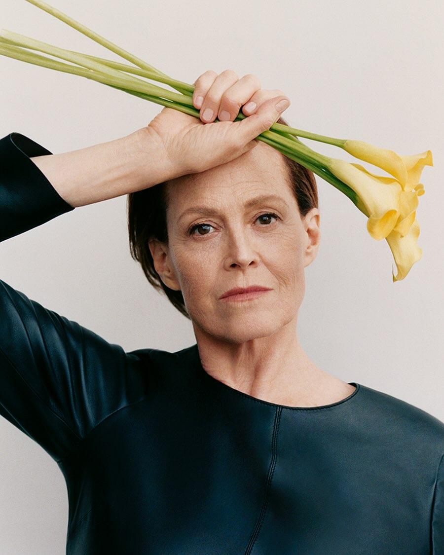 Headshot of adult woman in dark top holding three yellow Calla lilies overhead against plain backdrop, neutral studio lighting.