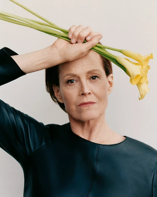 Headshot of adult woman in dark top holding three yellow Calla lilies overhead against plain backdrop, neutral studio lighting.