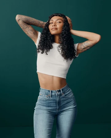 Woman with curly dark hair and arm tattoos wearing a white cropped t-shirt and blue jeans against a dark green backdrop