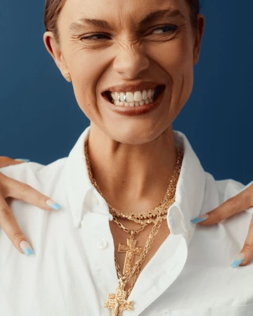 Close-up of a smiling adult woman with a playful expression, wearing a white shirt and gold necklaces against a blue background.
