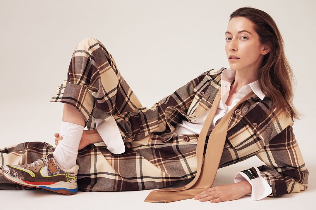 Adult female posing in a brown and tan plaid outfit with a white shirt and brown tie, lying on a white surface, looking at the camera.