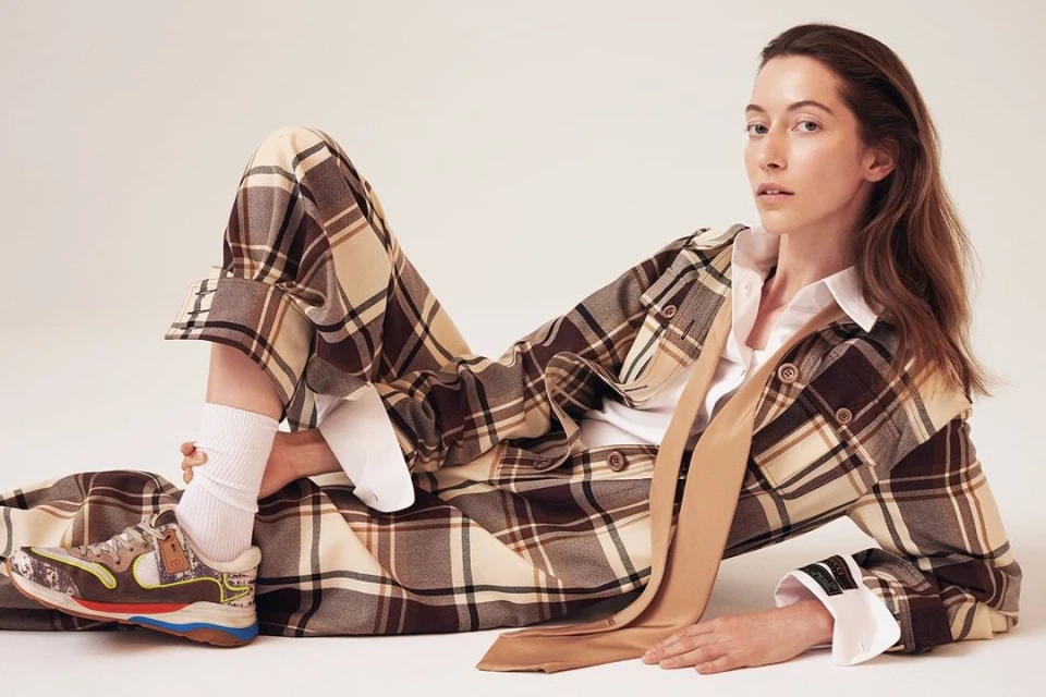 Adult female posing in a brown and tan plaid outfit with a white shirt and brown tie, lying on a white surface, looking at the camera.