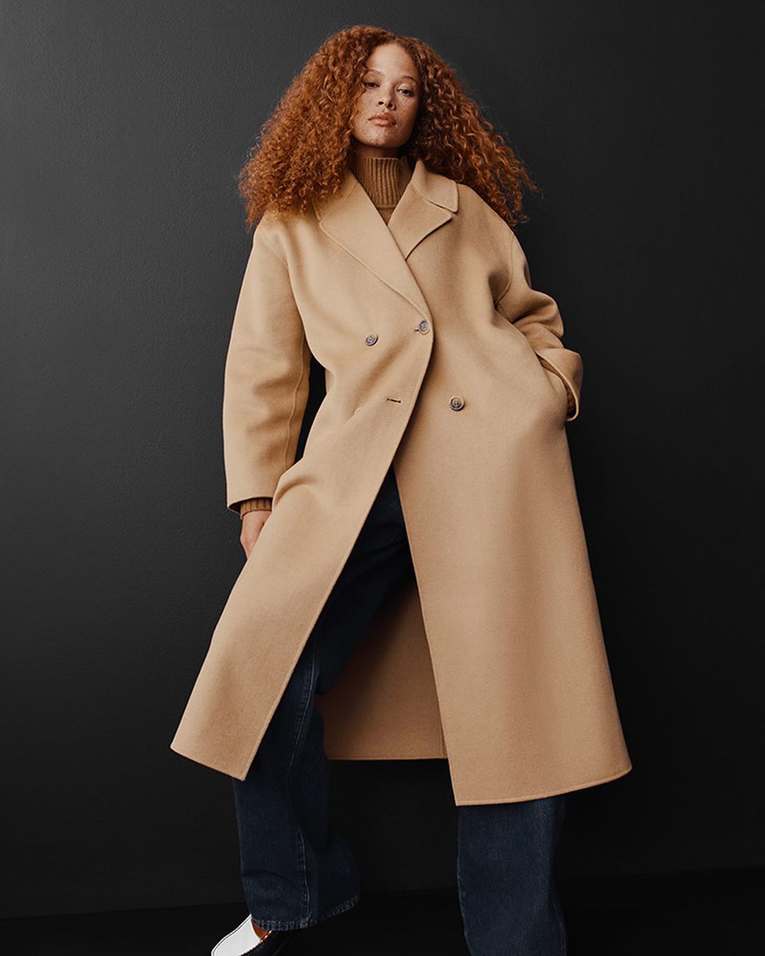 A red-haired woman poses in a tan double-breasted coat, dark blue jeans, and white shoes against a dark background.
