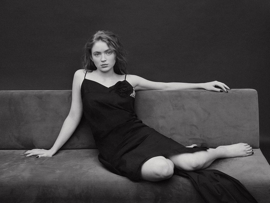 Sadie Sink in a black slip dress reclining on a grey upholstered sofa against a dark studio backdrop, photographed by Bjorn Iooss.