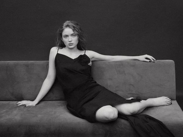 Sadie Sink in a black slip dress reclining on a grey upholstered sofa against a dark studio backdrop, photographed by Bjorn Iooss.