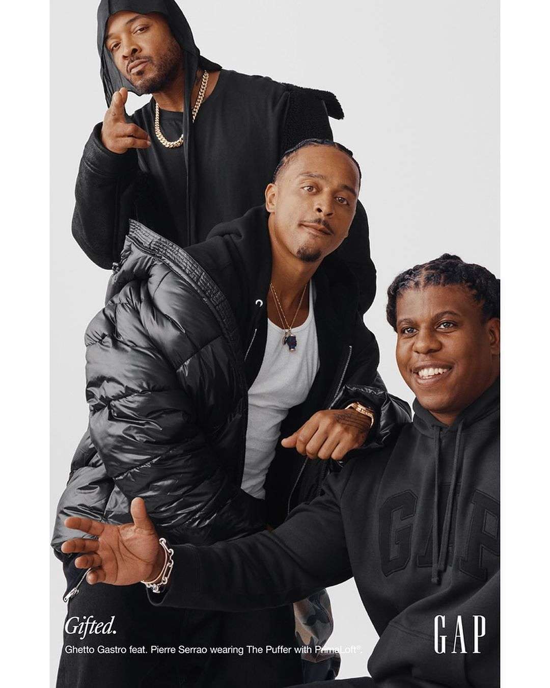 Three men of African descent pose indoors for a fashion advertisement in black hoodies and a puffer jacket against a white backdrop. 