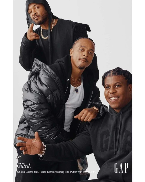 Three men of African descent pose indoors for a fashion advertisement in black hoodies and a puffer jacket against a white backdrop. 