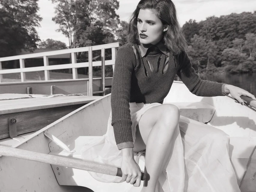 A young adult woman rows a boat on a lake; wearing a cut-out sweater and skirt, she exudes a blend of fashion and classic outdoor style.