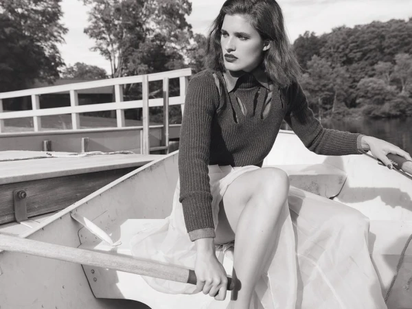 A young adult woman rows a boat on a lake; wearing a cut-out sweater and skirt, she exudes a blend of fashion and classic outdoor style.