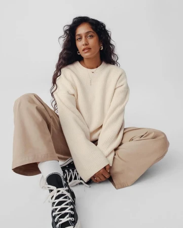 Woman with long dark hair wearing a cream sweater and beige trousers sitting on a white background