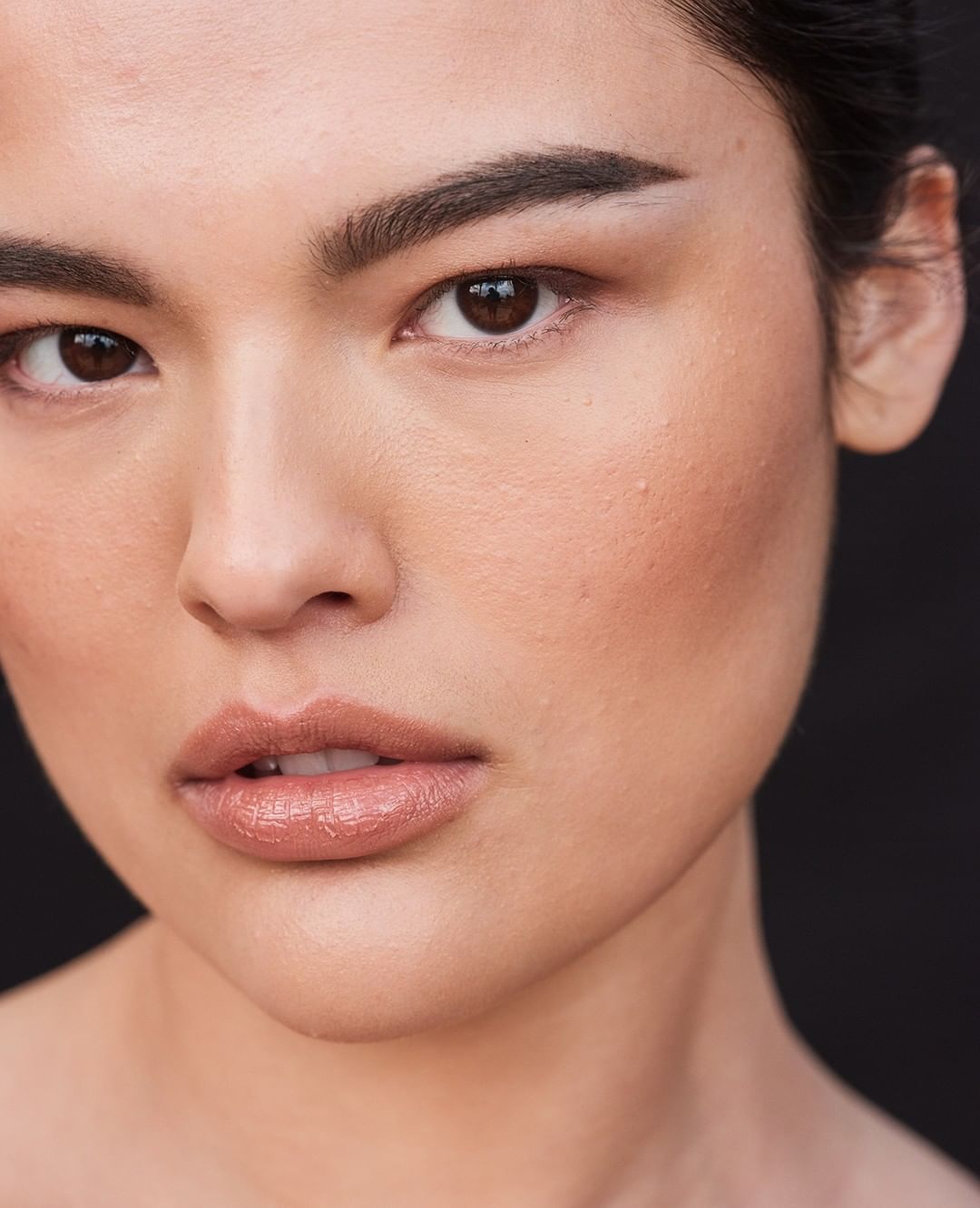 Close-up portrait of a young adult woman with dark hair, featuring soft, natural makeup, and a dark background, highlighting her facial features.