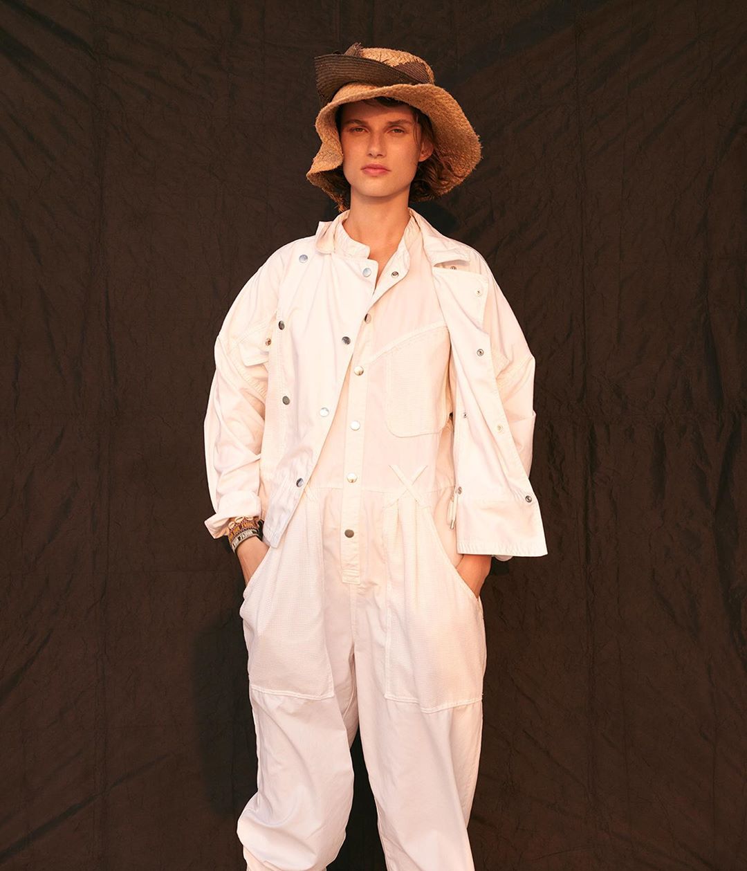 Woman poses in white coveralls and straw hat against a dark backdrop. Fashion shot with neutral tones and casual style in a studio setting.