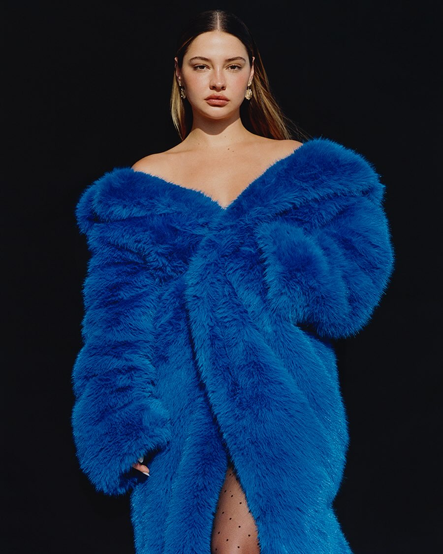A woman in a bright blue faux fur coat poses against a black background, looking directly at the camera in a fashion-style portrait.