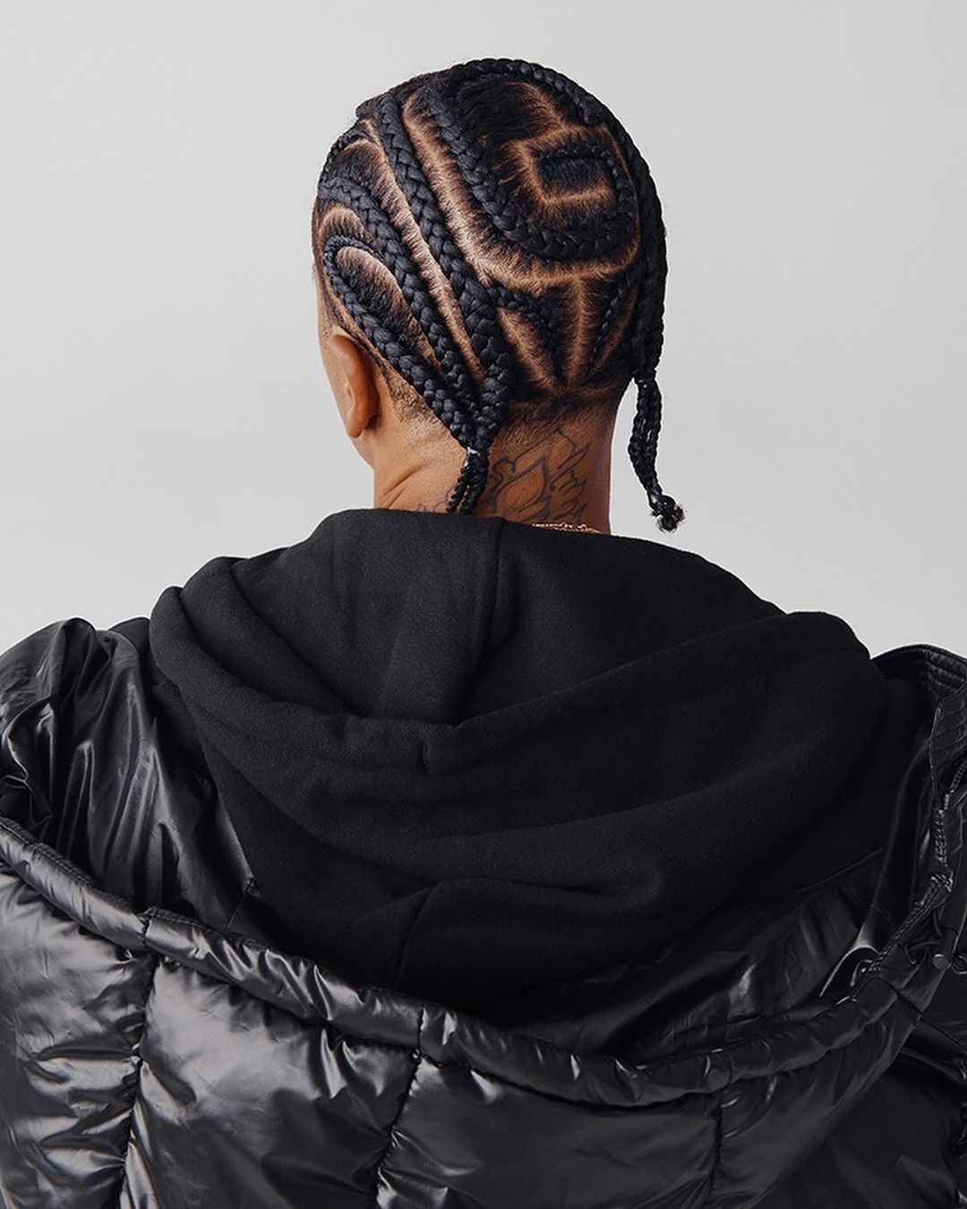 Young adult viewed from the back shows braided hair with an interesting design, hooded black sweatshirt, and puffy black jacket.