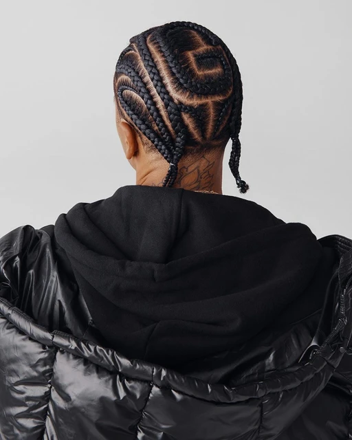 Young adult viewed from the back shows braided hair with an interesting design, hooded black sweatshirt, and puffy black jacket.