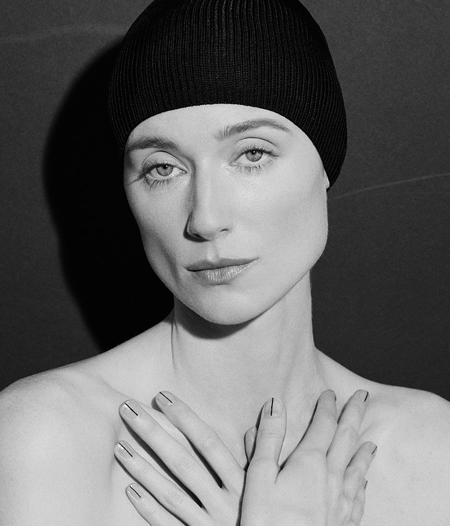 Black and white portrait of a woman with a black beanie, her hands crossed in front of her chest, posing against a dark background.