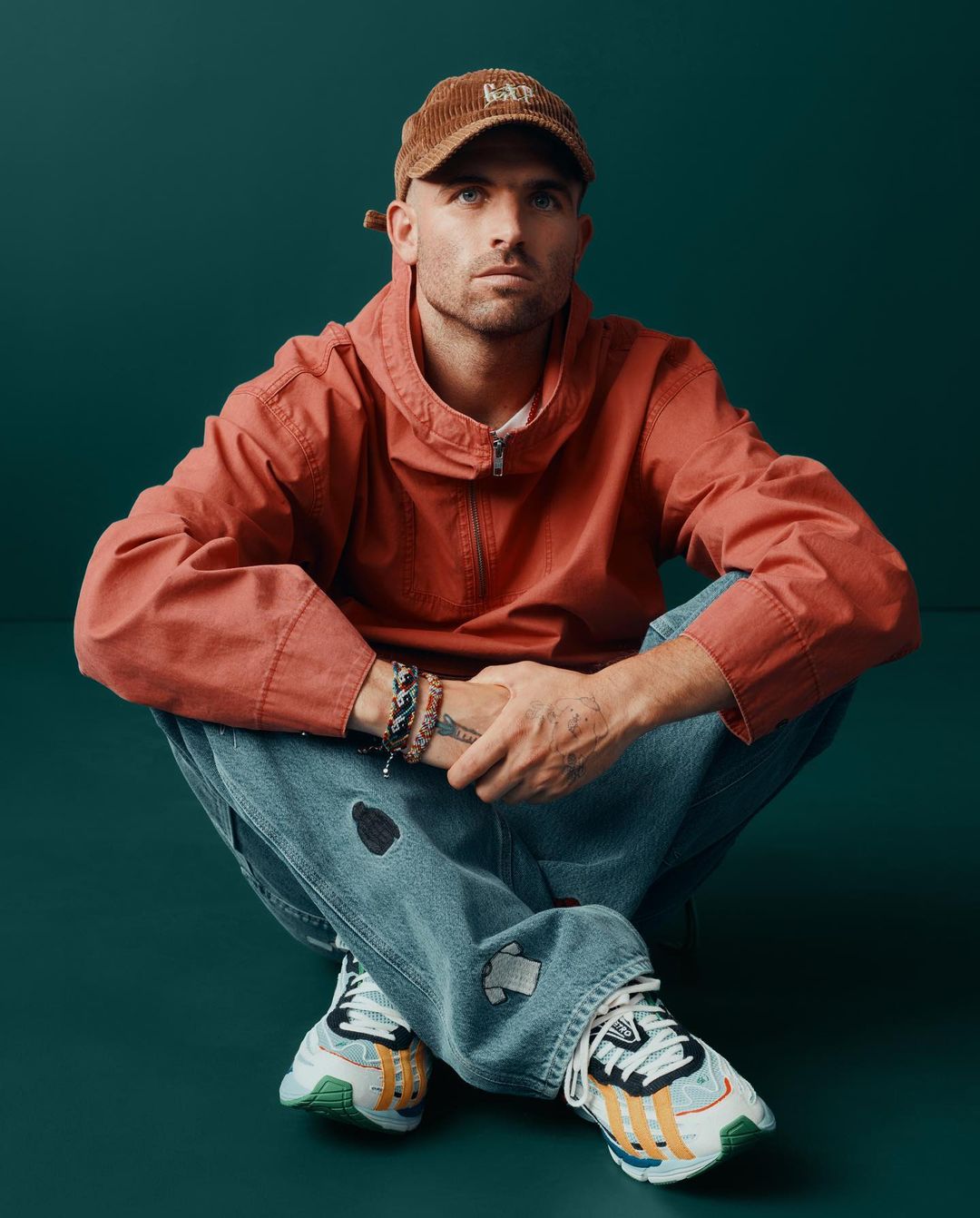 Young adult male in a red jacket and cap sits cross-legged on a green surface, looking directly at the camera in an eye-level shot.