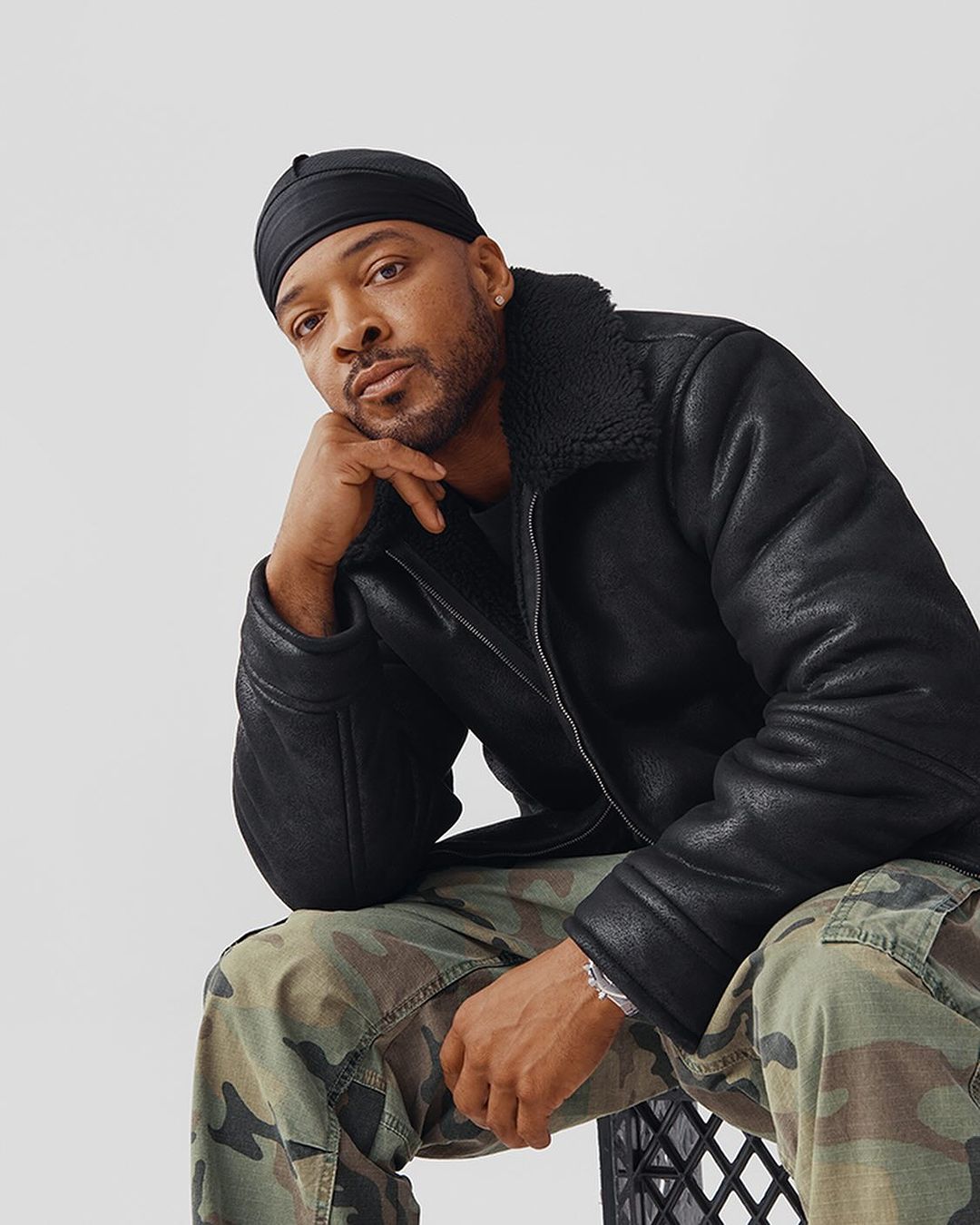 Stylish adult male poses indoors wearing a black shearling jacket, camouflage pants, and a durag, creating a fashionable portrait.